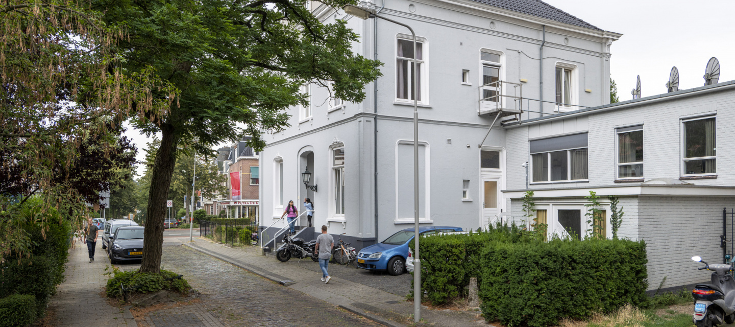 wit gebouw met grijs dak met geparkeerde auto's, straat en bomen ervoor