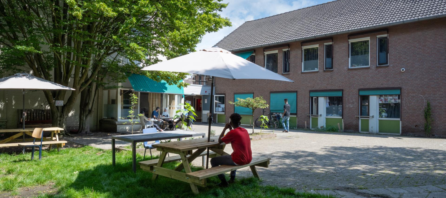 jongen met rood shirt zit buiten op een bankje achter het azc