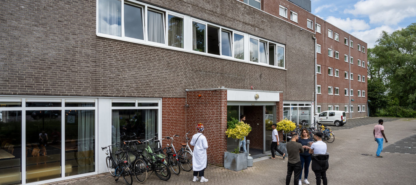 Flatgebouw voor opvang asielzoekers. Er staan wat mensen voor de ingang te praten.
