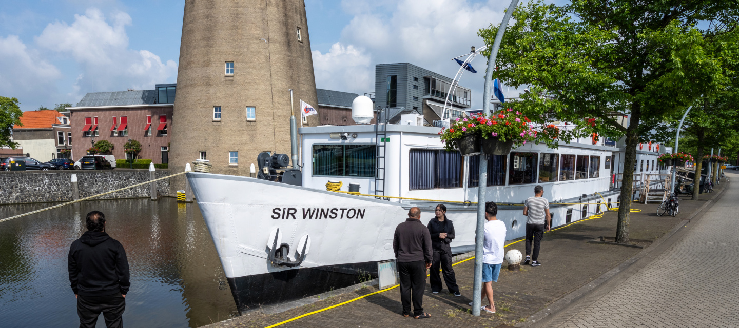 Asielzoekers kijken vanaf de kade naar het schip waar zij wonen. Op de achtergrond staat een molen.