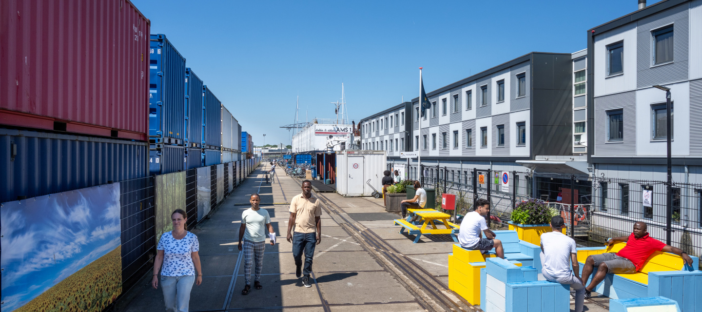 Medewerkers en statushouders lopen zitten buiten op de kade naast het drijvend ponton met woonblokken.