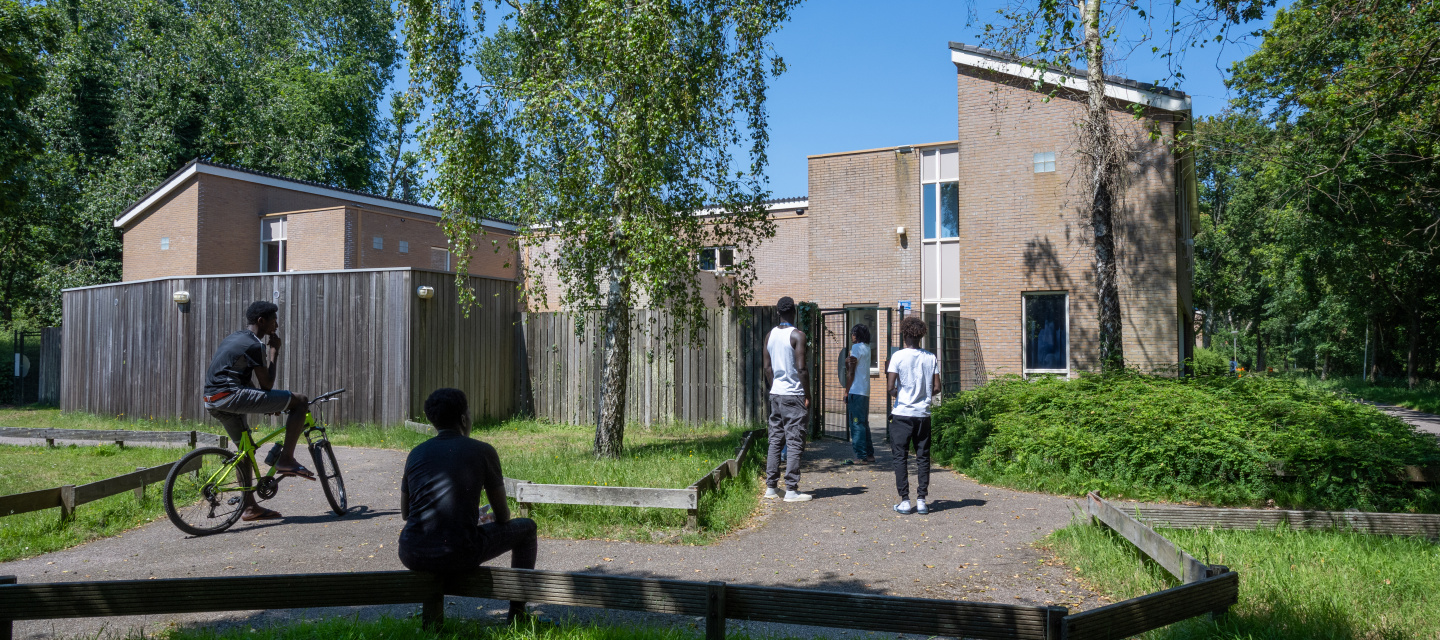 Minderjarige jongens staan en zitten voor de ingang van hun opvanglocatie in een groene omgeving.