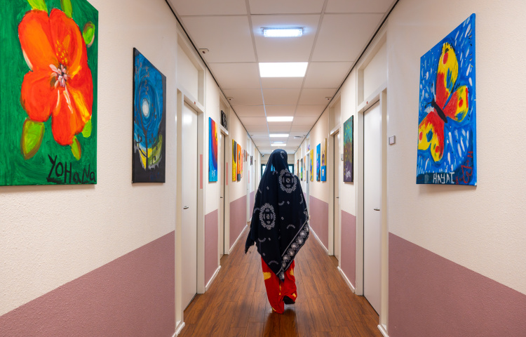 Jonge vrouwelijke asielzoeker in jurk loopt door de gang in het azc, we zien haar op de rug, er hangen kleurrijke schilderijen aan de muur