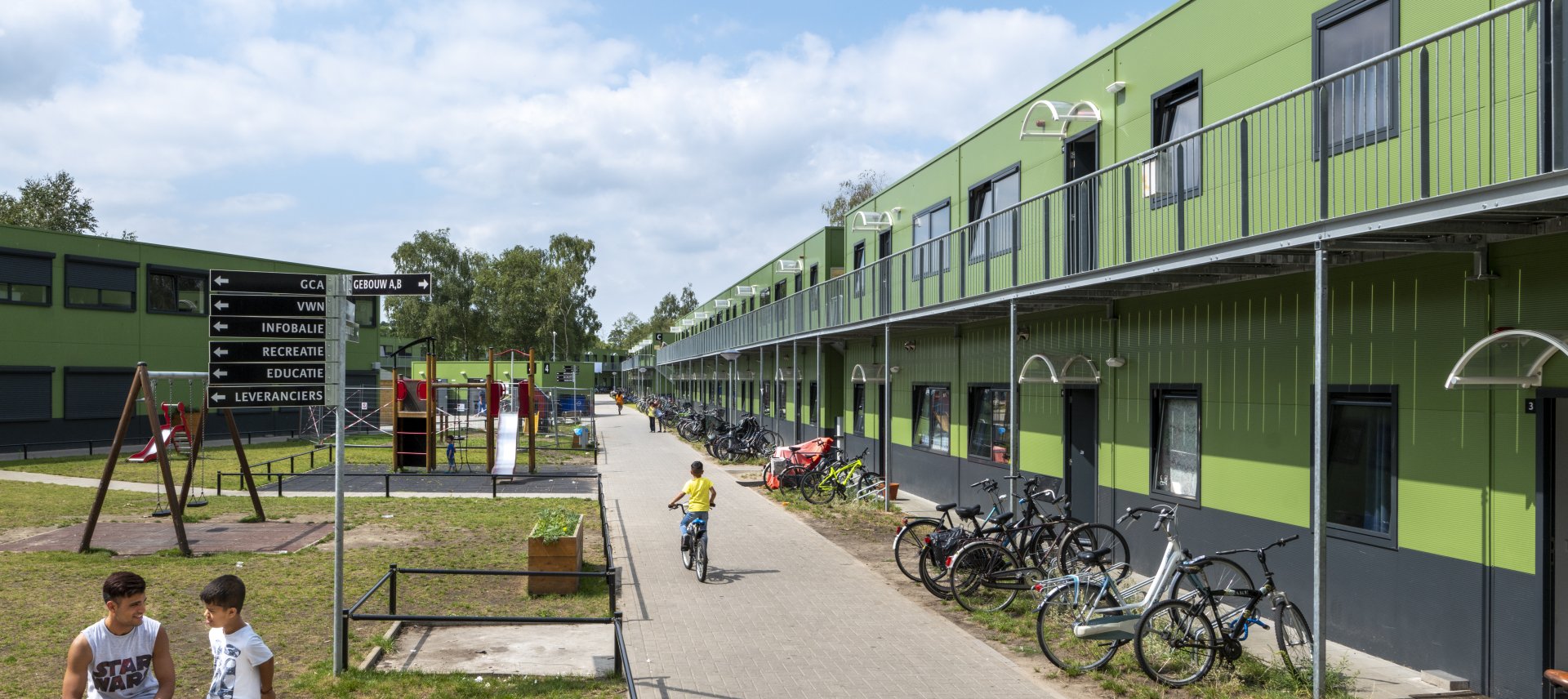 Groene gebouwen met twee verdiepingen, fietsen, gras en speeltuin met kinderen ervoor