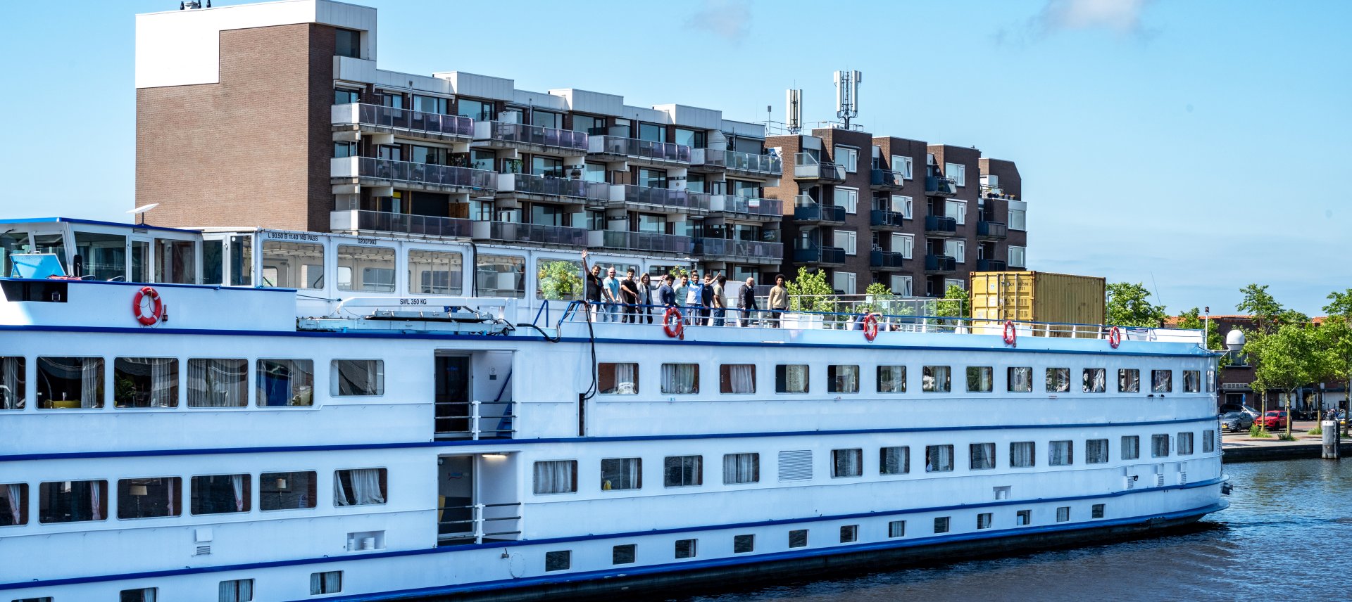 Asielboot ligt aan de kade, op het dek staan mensen te zwaaien.