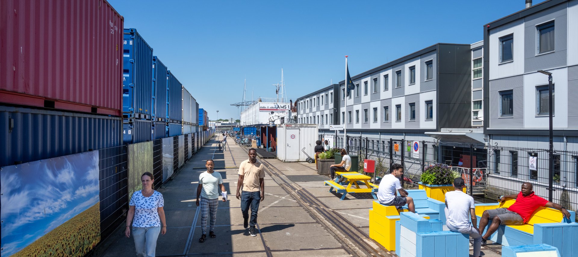 Medewerkers en statushouders lopen zitten buiten op de kade naast het drijvend ponton met woonblokken.