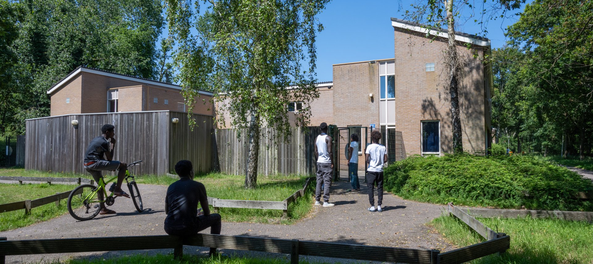 Minderjarige jongens staan en zitten voor de ingang van hun opvanglocatie in een groene omgeving.