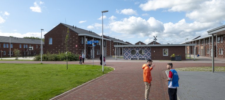 2 pratende mensen staan op een terrein met daarachter gebouwen van twee verdiepingen met puntdak