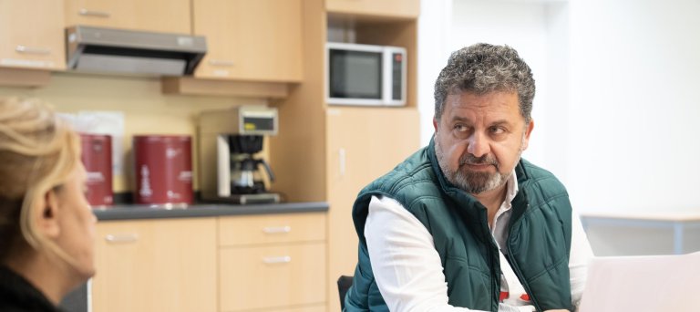 Man met groene bodywarmer en wit shirt zit aan een keukentafel, en heeft papier in de hand. Hij kijkt een blonde vrouw die van achteren gefotografeerd is aan. 