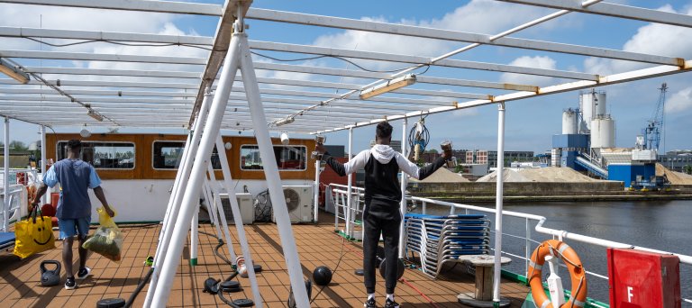 Twee mannen zijn aan het fitnessen op het dek van een asielboot.