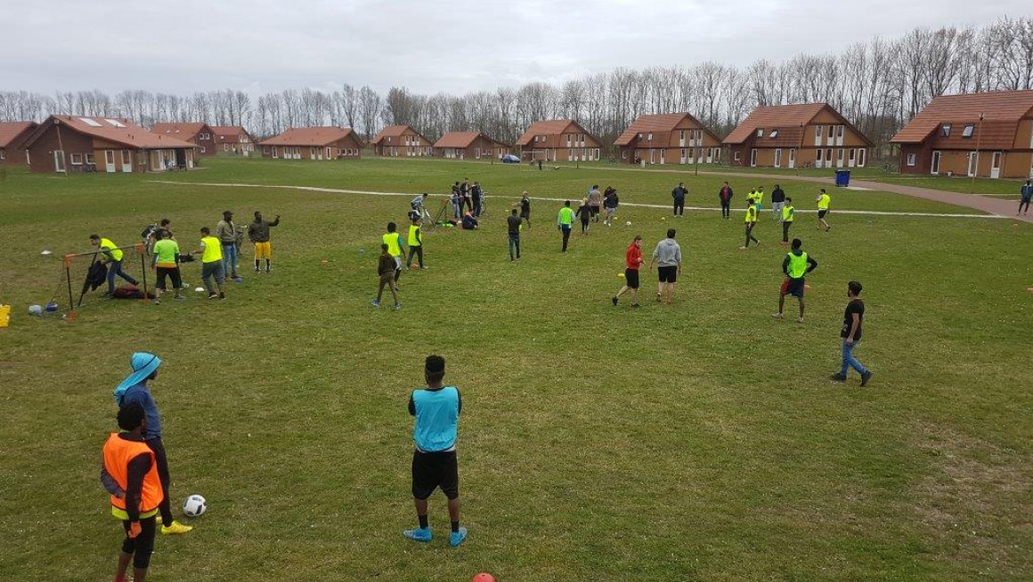 groot grasveld met voetballende bewoners en op de achtergrond woongebouwen met daarachter bomen