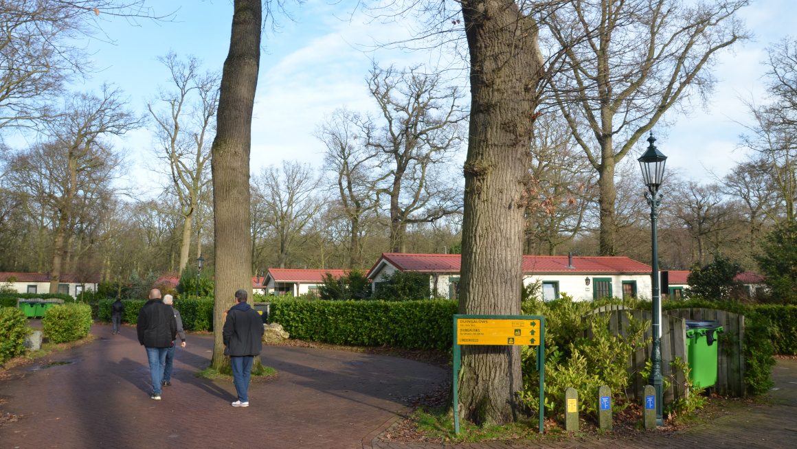 witte woonunits met rode daken en her en der grote bomen en heggen, op de voorgrond lopen 3 mannen