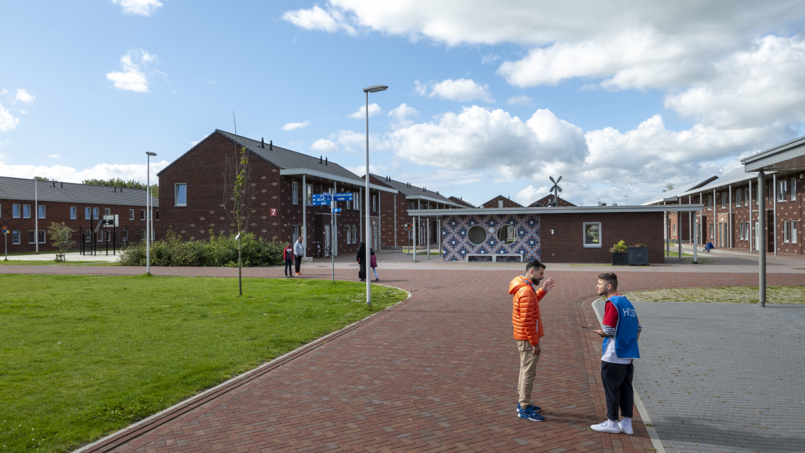 2 pratende mensen staan op een terrein met daarachter gebouwen van twee verdiepingen met puntdak