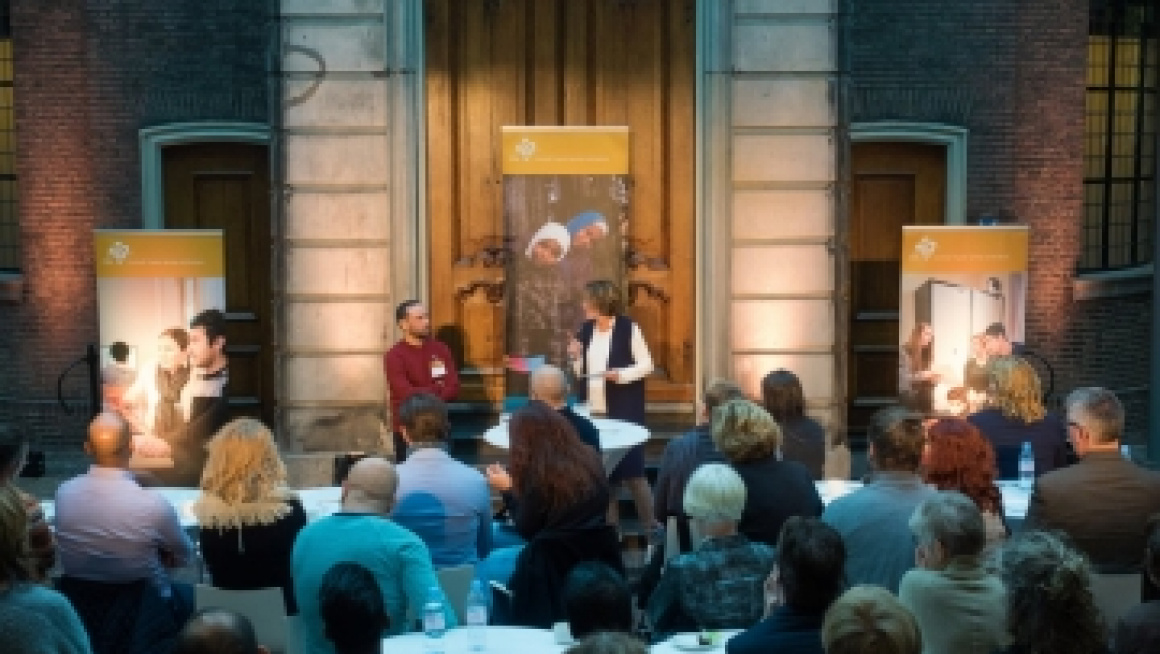 vrouw staat op een podium met microfoon in hand en praat met een man in een stoel