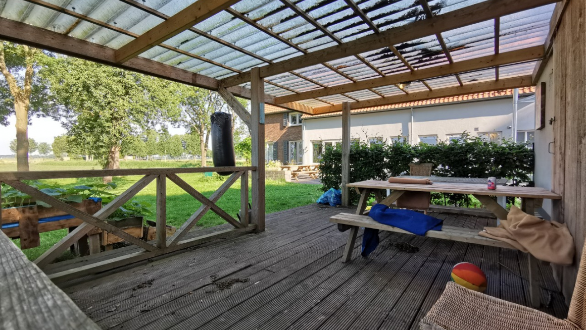 houten veranda met picknicktafel en overkapping, daarvoor gras en bomen
