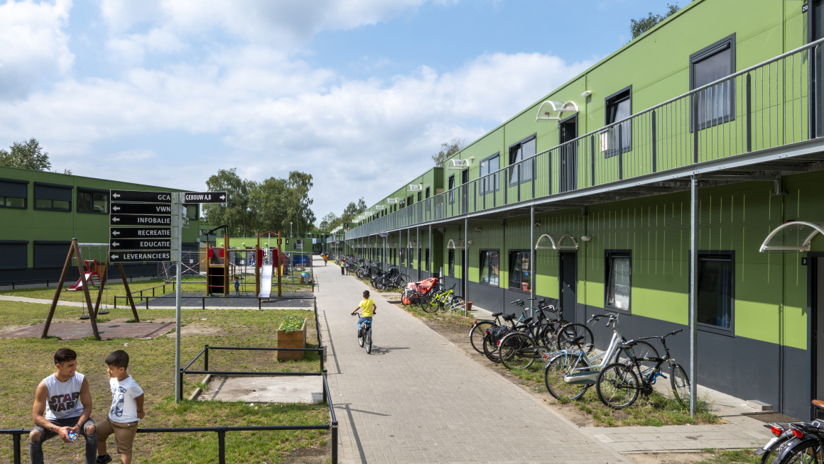 Groene gebouwen met twee verdiepingen, fietsen, gras en speeltuin met kinderen ervoor