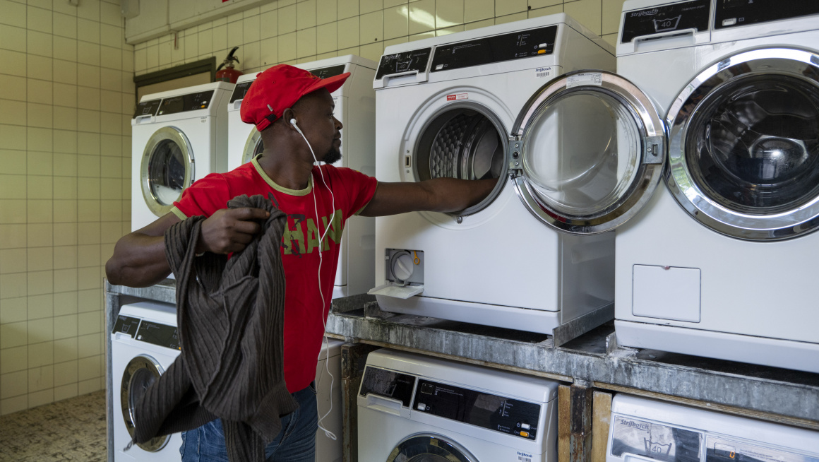 wasserette met tegels op de vloer en muur en 8 wasmachines/drogers en een bewoner met rood t-shirt en rode pet die was in de wasmachine doet