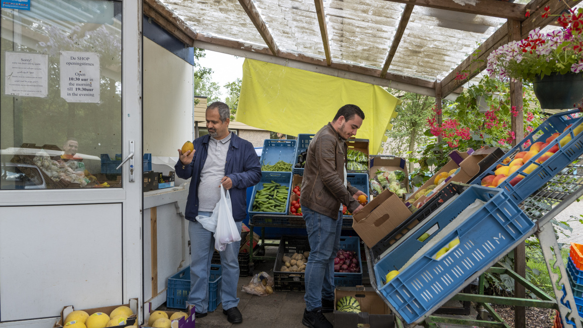 groente-/fruitkraam met plastic overkapping en stellingen met blauwe bakken met fruit en groenten en twee mannen die fruit pakken