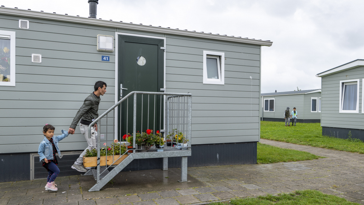 Grijze stacaravans met witte kozijnen en puntdak, bewoner met kind lopen trapje met bloembakken met bloemen op naar ingang stacaravan