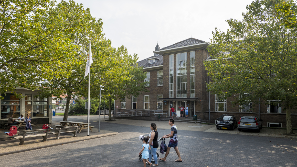 binnenplaats met parkeerplaatsen, het hoofdgebouw met bomen ervoor en op de voorgrond een man en vrouw met kinderwagen en kind aan de hand die het plein oversteken