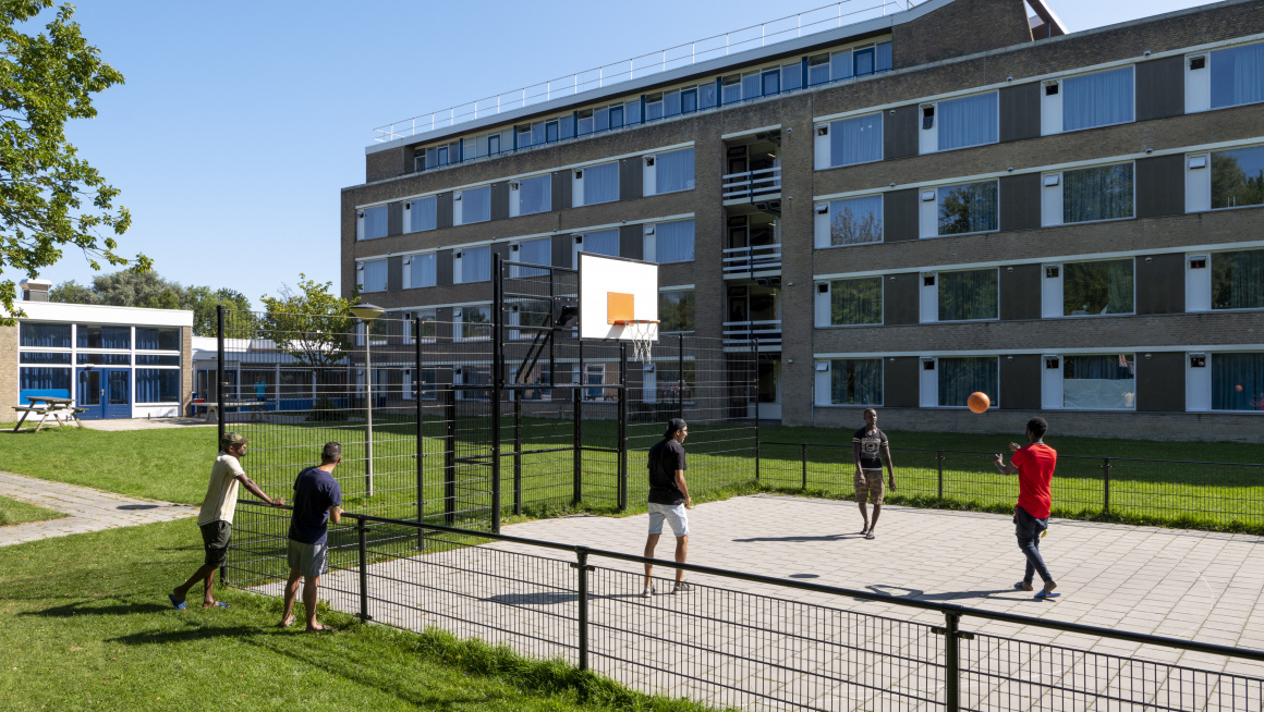 grasveld met betegeld basketbalveld met basketballende bewoners en op de achtergrond het woongebouw