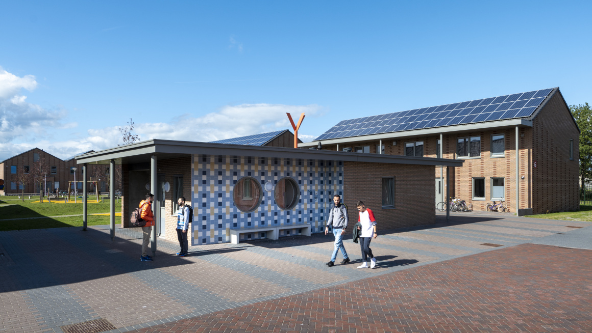 woongebouwen met twee verdiepingen met houten buitenkant en puntdaken met zonnepanelen, daarvoor een laag gebouw met plat dak en aan de buitenkant hout en wit met blauw en gele tegels, daarachter gras en rondom tegels waar vier bewoners lopen