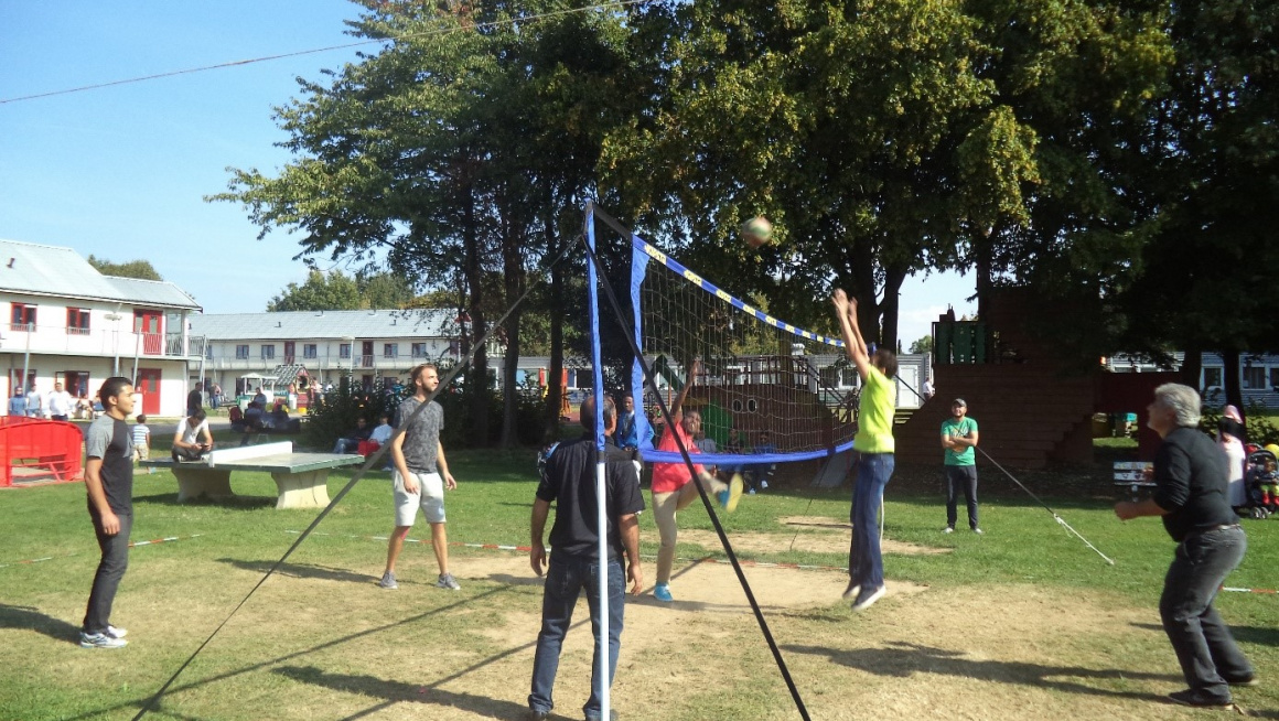 volleybalveld met spelende bewoners en op de achtergrond een tafeltennistafel, gebouwen en bomen