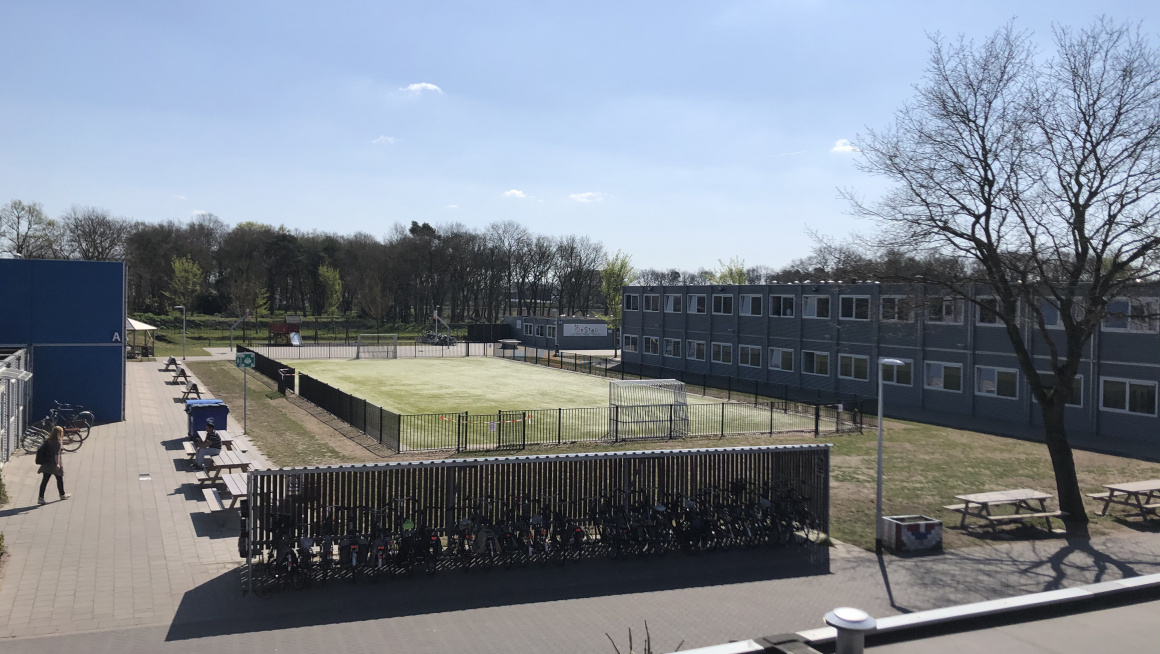 bovenaanzicht sportveld met gebouwen, bomen en een fietsenstsalling met fietsen