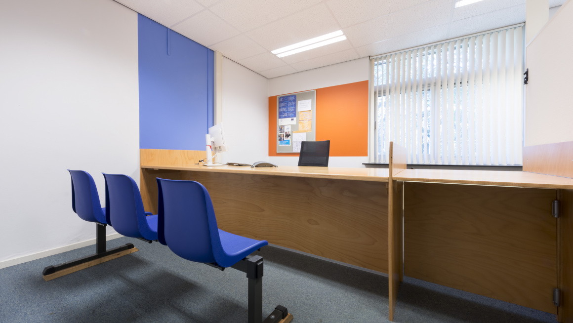 spreekkamer met op de voorgrond 3 blauwe stoelen, daarachter een bureau en daaracher een raam met witte lamellen, de muren zijn wit, blauw en oranje