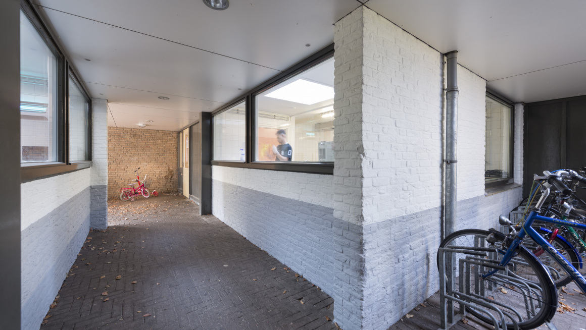 Buitenkant woning met wit/grijze muur en zwarte kozijnen, een bewoner achter het raam en rechts voor het gebouw fietsen in een fietsenrekmet fietsen