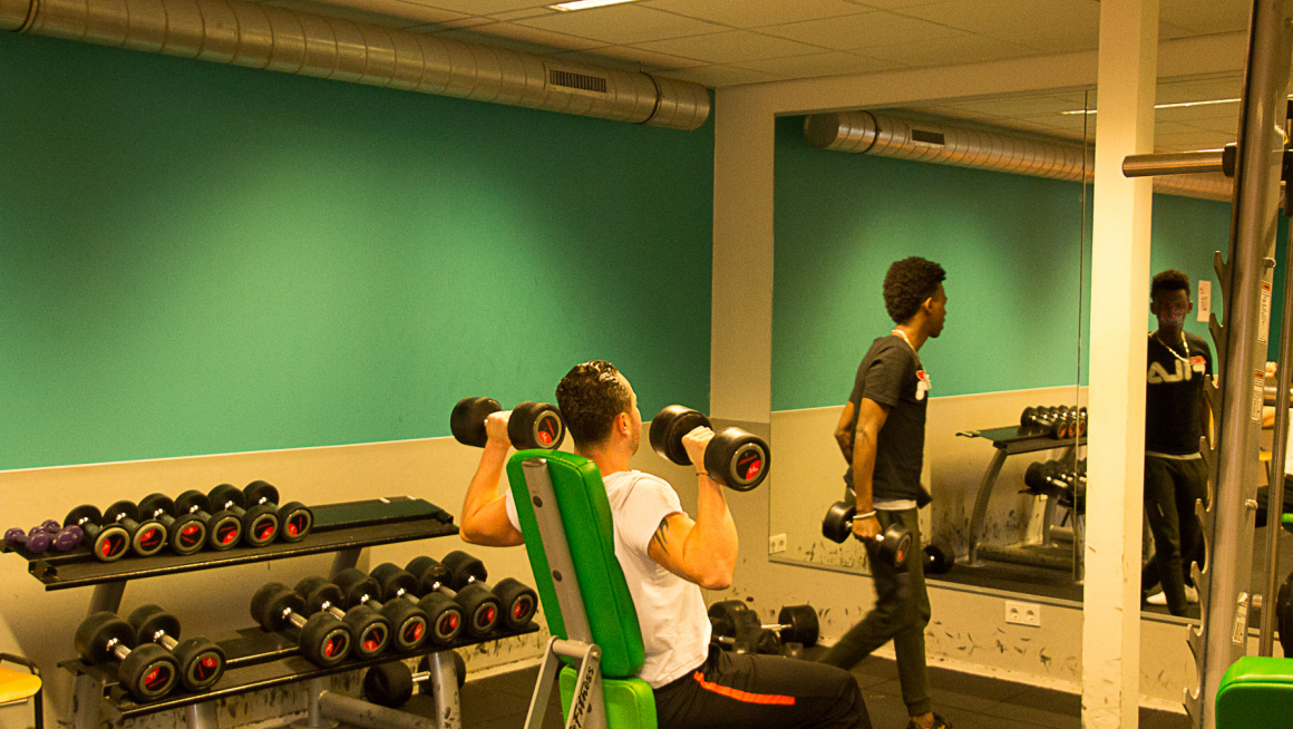 fitnessruimte met grote spiegel, groene muur, gewichten en twee trainende mannen