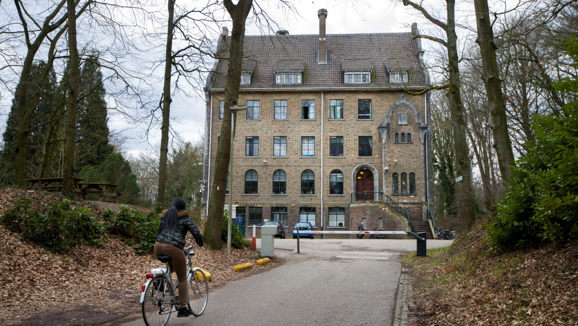 voorgevel van gebouw met vijf verdiepingen daarvoor een slagboom en een weg met aan beide kanten bomen en struiken en op de weg een fietsende vrouw