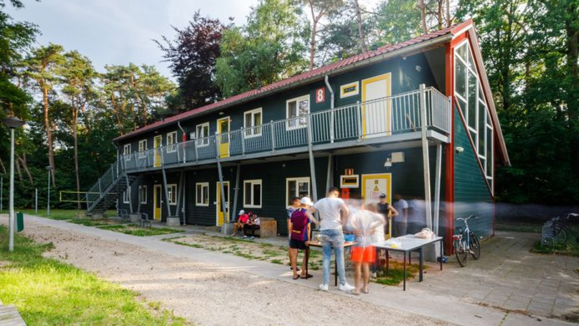 groen woongebouw met twee verdiepingen, rood dak en gele deuren daarvoor gras en een betegeld pad met een groepje bewoners in gesprek