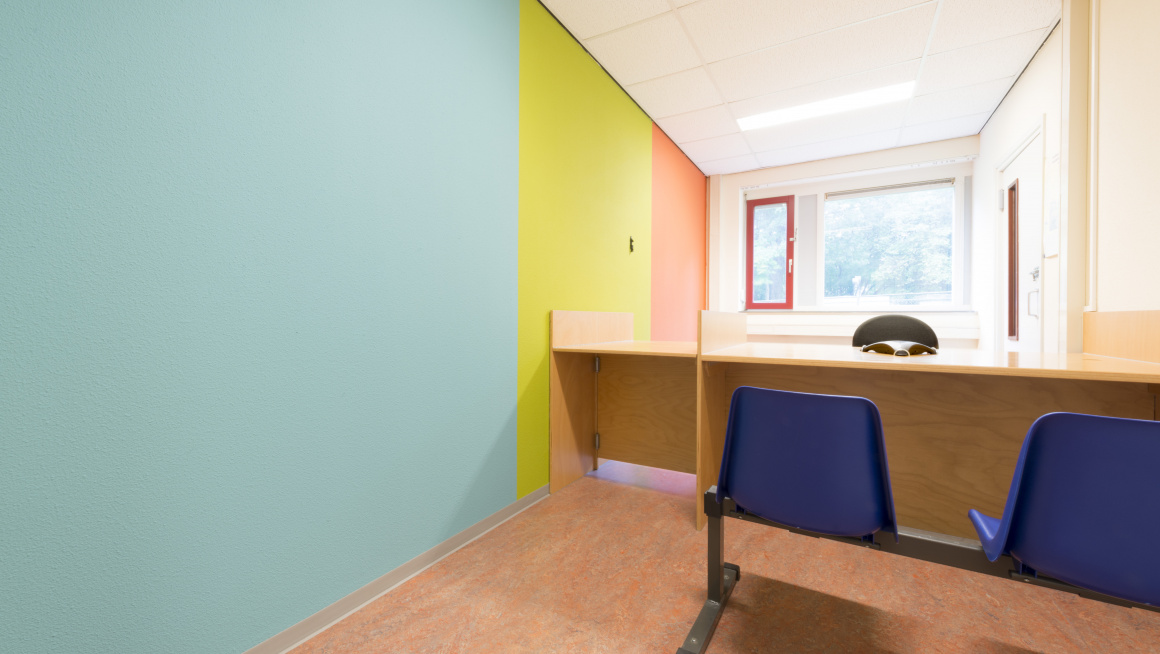 Spreekkamer met muur in blauw, geel en oranje, raam, bureau en blauwe stoelen