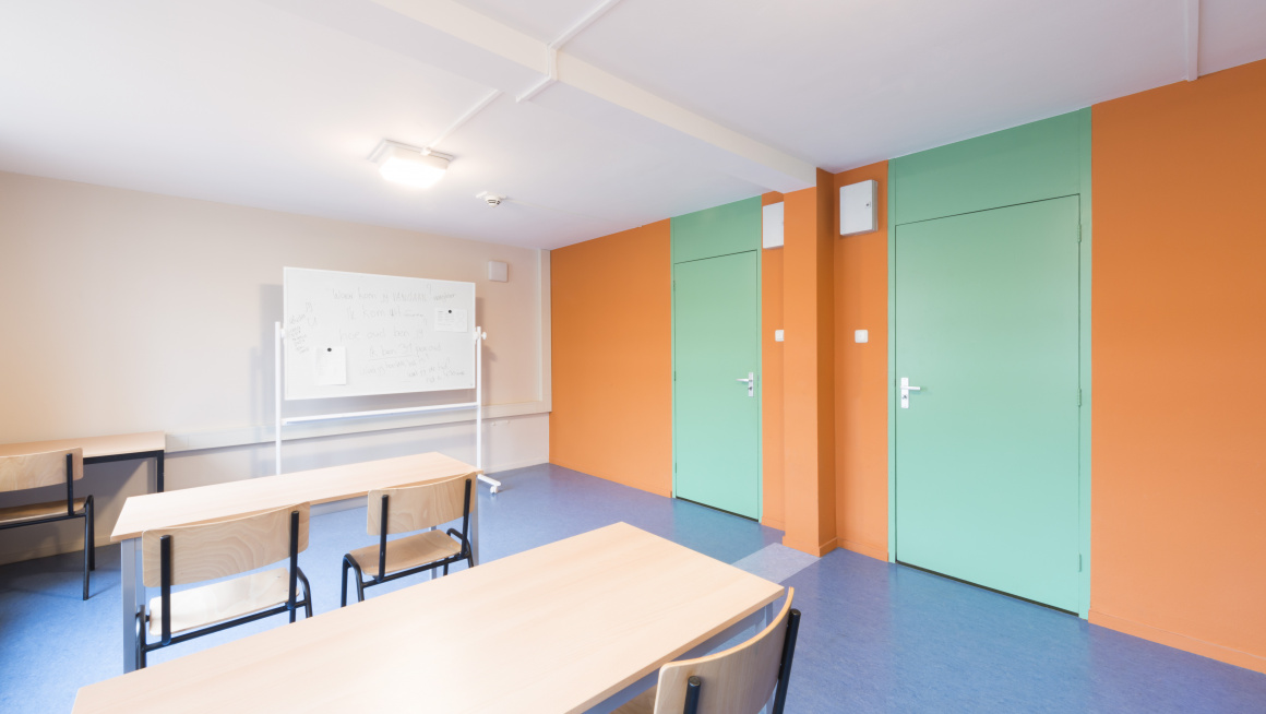 Leslokaal met witte en oranje muren en mintgroen deuren, tafels en stoelen en een whiteboard