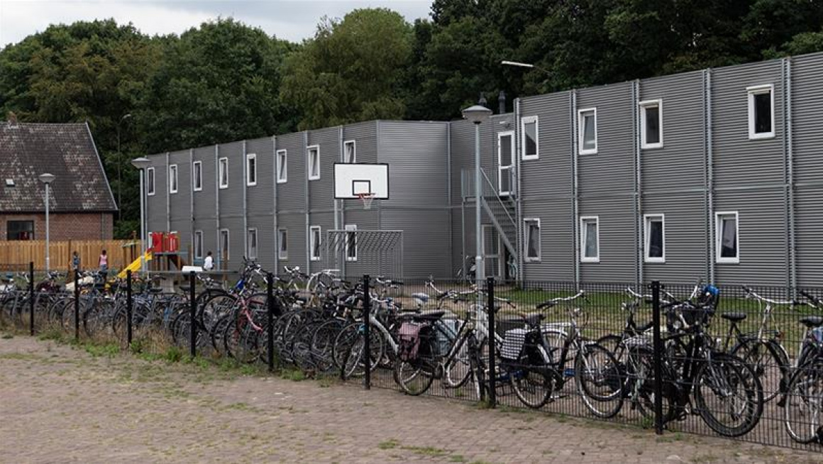 grijze woonunits met basket, speeltoestel en fietsen ervoor en bomen erachter