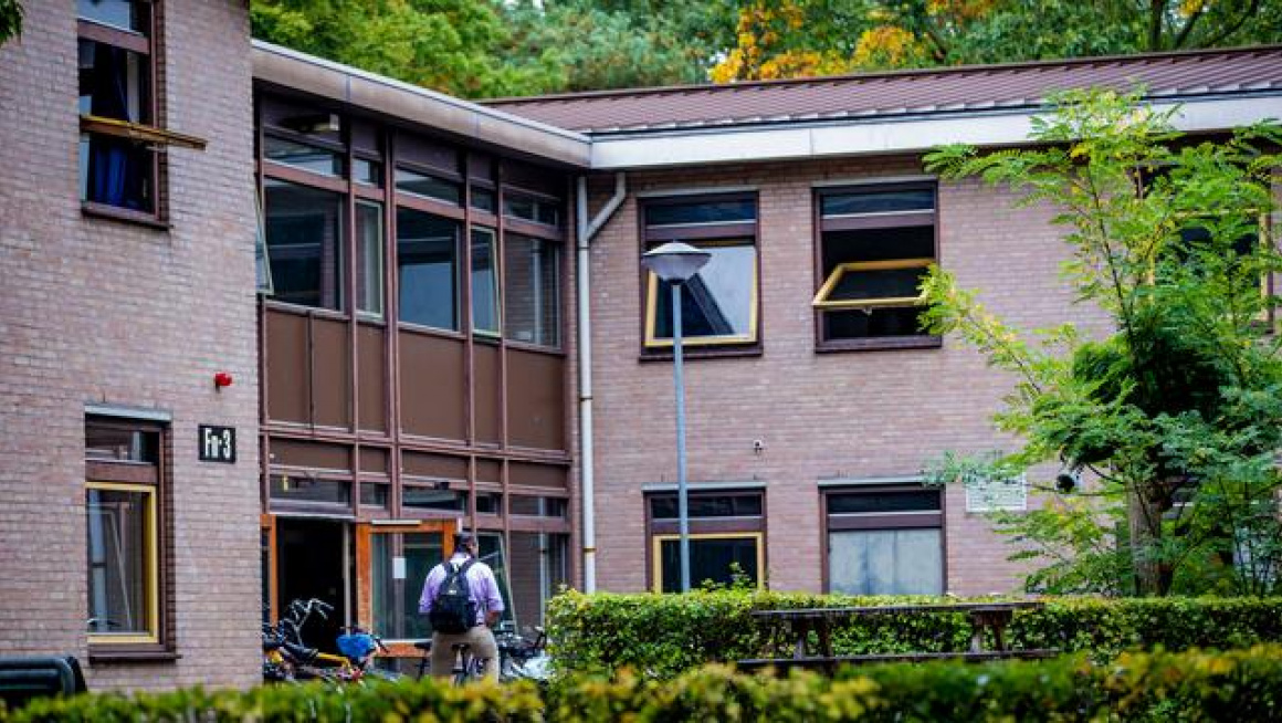 buitenkant gebouw met daarvoor een boom, struiken, een bewoner op de fiets en een lantaarnpaal