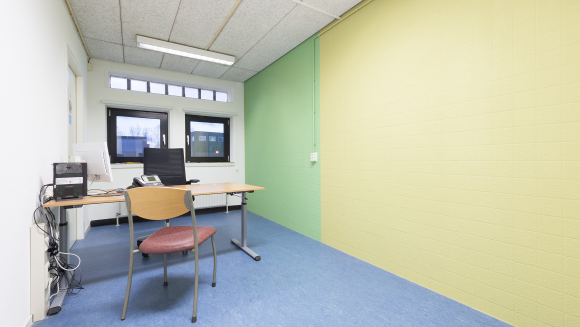 kantoorruimte met bureau, bureaustoel en gewone stoel ervoor. Blauwe vloer en witte, groene en gele muren en twee ramen