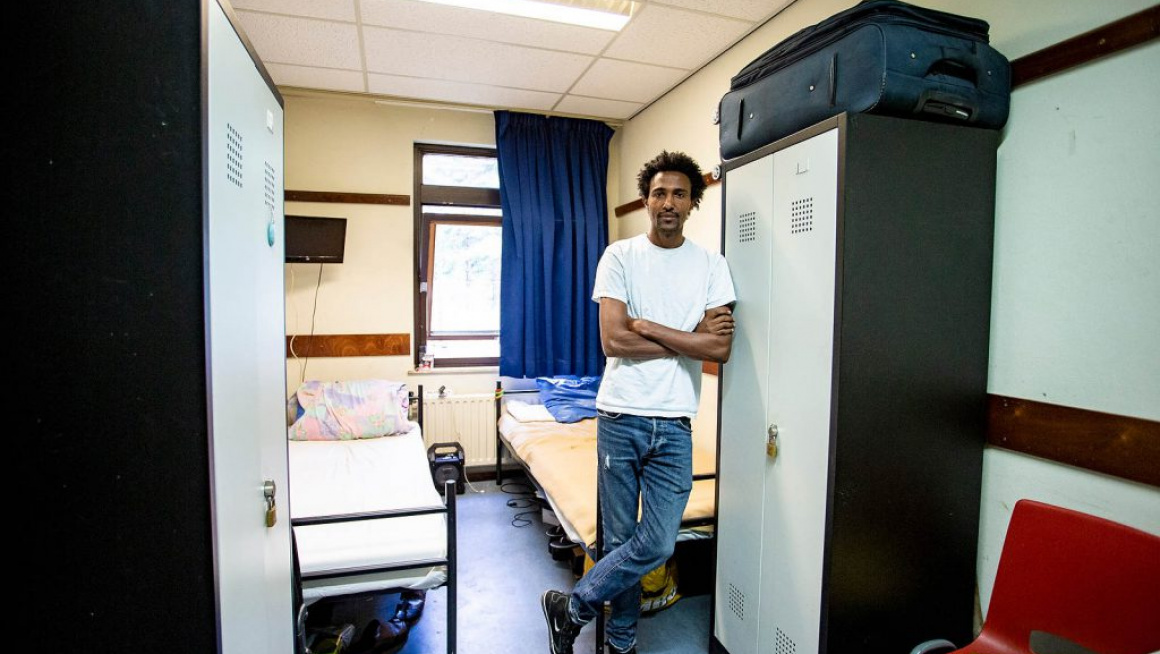 jonge man op bewonerskamer met twee bedden, twee kasten, rode stoel en blauwe gordijnen