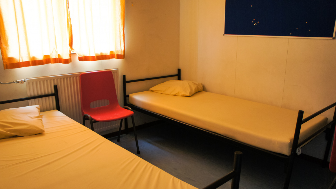 bewonerskamer met twee bedden en een rode stoel ertussen, daarachter een raam met dichte oranje gordijnen