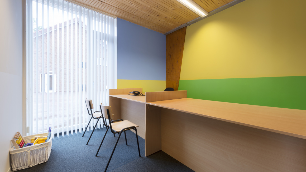 spreekkamer met 2 stoelen voor bureau, groot raam, geel blauwe en geel groene muur en een bak met speelgoed