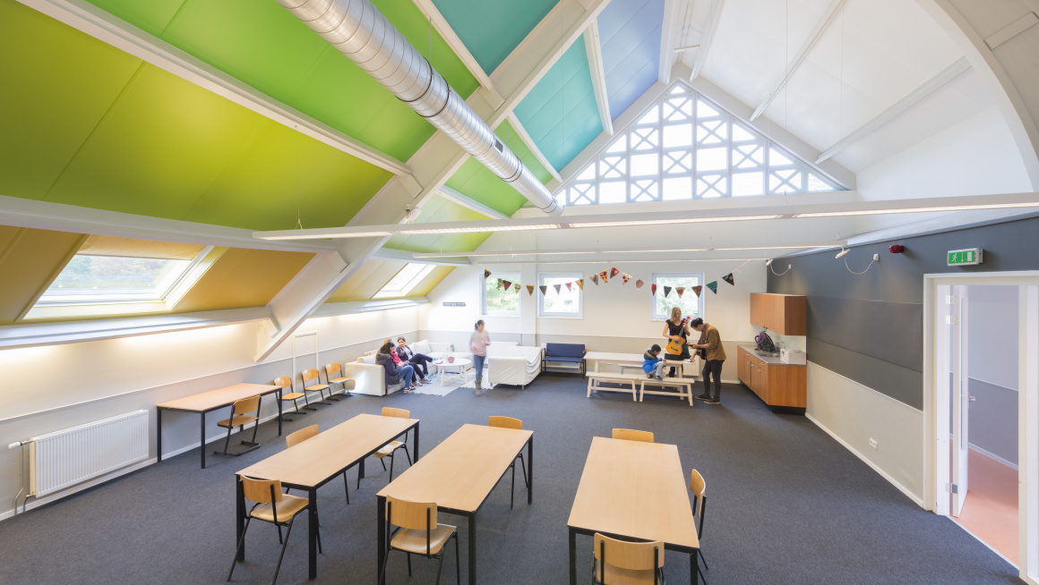 recreatieruimte met tafels en stoelen, keukentje, witte banken en op het schuine plafond verschillende kleuren (wit, blauw, turqoise, groen, geel en oranje)