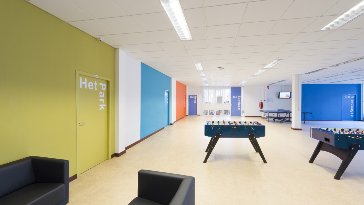 Recreatiezaal met gekleurde muren in groen, wit, blauw en oranje, beige vloer, zware bankjes, twee voetbaltafels en een tafeltennistafel