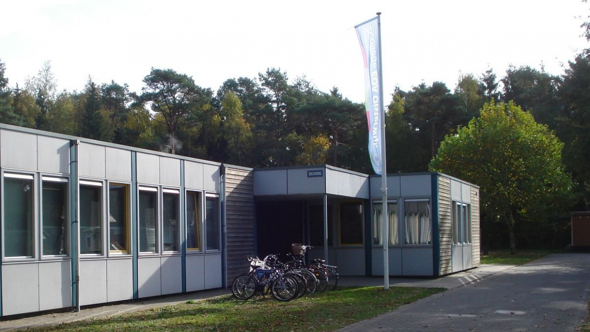 schoolgebouw, 1 verdieping, plat dak met vlaggenmast met vlag, fiestenrek met fietsen en gras ervoor