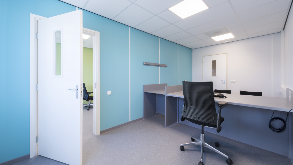 spreekkamer met grijze vloer en witte en blauwe muren, een grijs bureau en zwarte stoelen