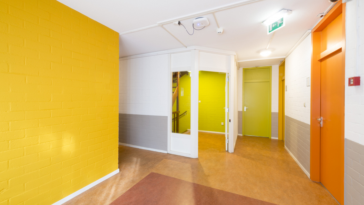 gang met gele vloer met oranje vlak, wit/grijze, groene en gele muren en deuren in oranje, groen en geel