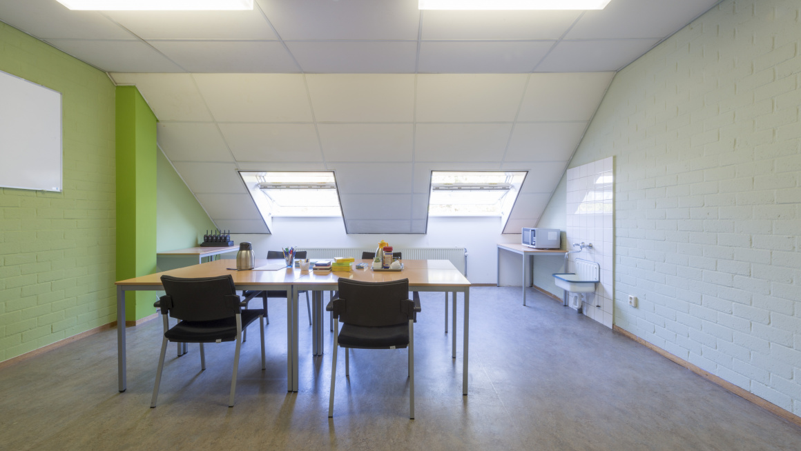 werkplek op zolder met vergadertafel en vier zwarte stoelen, een vloer met houtnerf witte muren en een groene muur met whiteboard en twee dakramen