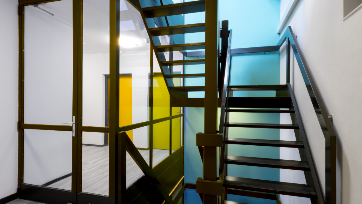 trappenhuis met zwarte trap, grijze vloer, blauwe en witte muren en een gele en groene deur