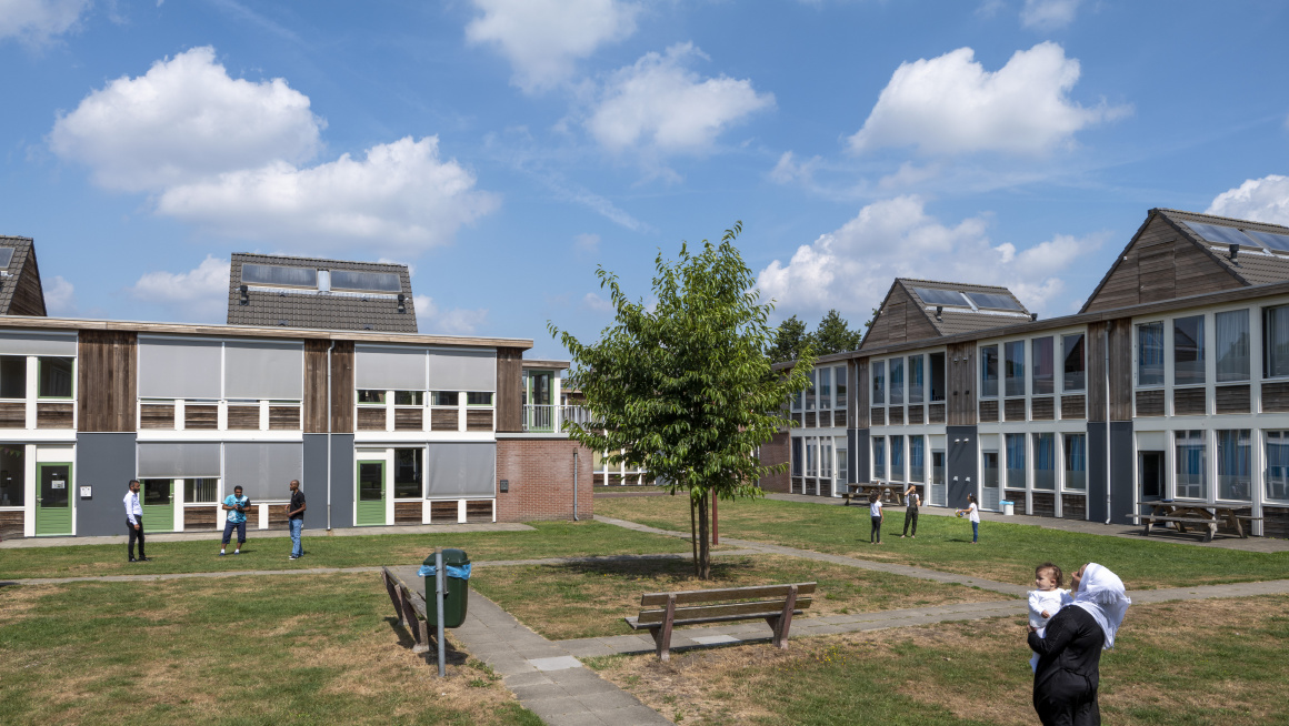 woongebouwen met aan de buitenkant hout en witte kozijnen, deels plat dak deels puntdak, daarvoor gras, een boom en paden en bewoners die praten, wandelen, spelen