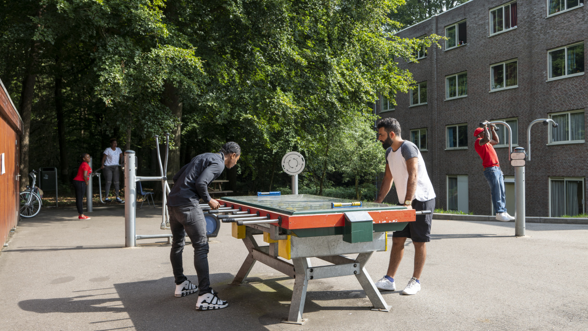 buitenterrein met betonnenvloer en daarop een voetbalspeltafel en fitnessapparatuur en bewoner die daar gebruik van maken. op de achtergrond een gebouw van vier verdiepingen met plat dak en daarnaast bomen